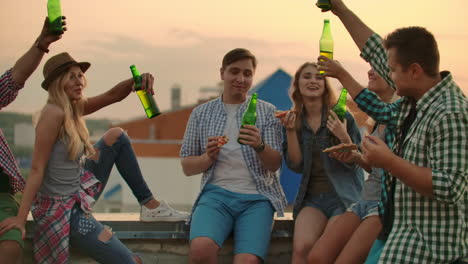 Three-girls-and-three-boys-in-plaid-shirts-drinks-beer-from-green-bottels-on-the-party-with-friends-on-the-roof-at-the-sunset.They-are-sitting-together-eat-hot-pizza-after-in-summer-everning.-A-man-says-a-toast-to-his-friend's-birthday.